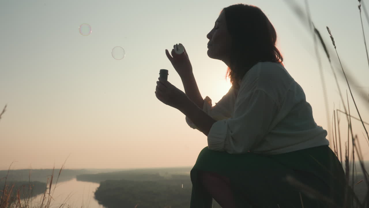 Silhouetted woman blowing bubble wand among tall grain stalks with glowing sun behind her, warm backlight illuminating soft breeze and floating bubbles over peaceful rural landscape at dusk