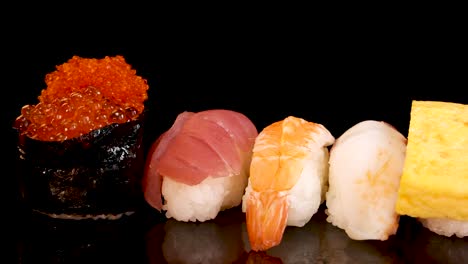 a variety of nigiri and sashimi pieces, including shrimp, tuna, salmon, egg, and roe, rotate smoothly on a glossy black surface under bright studio lighting