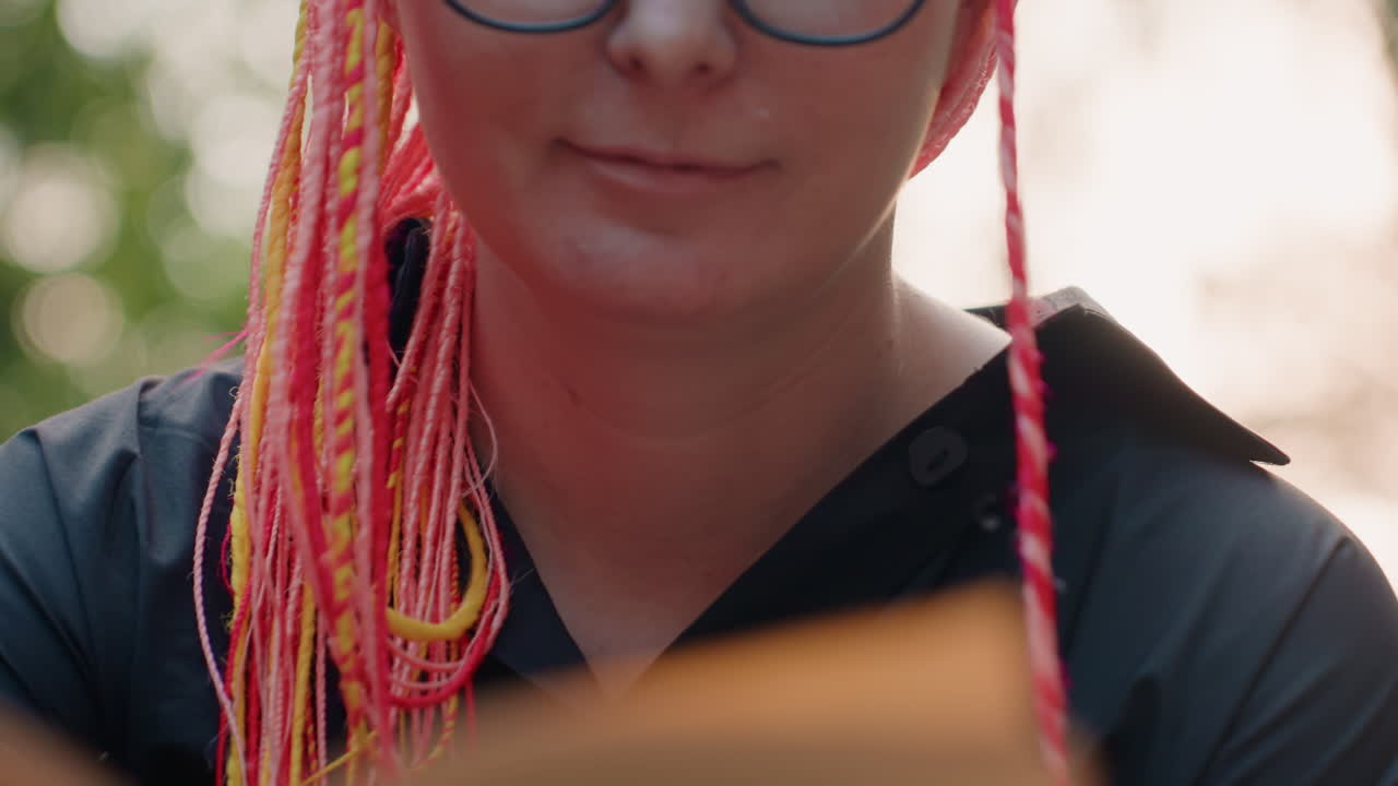 분홍색 땋은 머리를 한 소녀가 책에 몰두하고 있어요. 땋은 분홍색 머리를 한 여성이 얼굴 가까이에서 책을 열심히 읽고 있으며, 다채로운 땋은 머리를 한 젊은 여성이 평화로운 야외 환경에서 페이지에 집중하고 있어요.