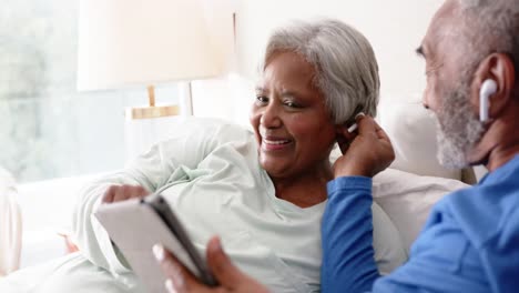 Feliz-Pareja-Birracial-De-Ancianos-Acostada-En-La-Cama,-Usando-Tableta-Y-Auriculares,-Inalterada,-En-Cámara-Lenta