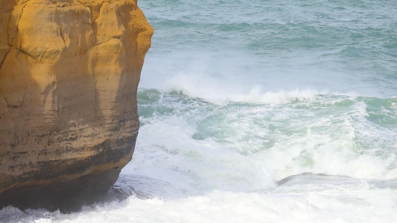 Dynamic ocean waves crash against rugged cliffs at Loch Ard Gorge, showcasing nature's power. Bright daylight illuminates the scene