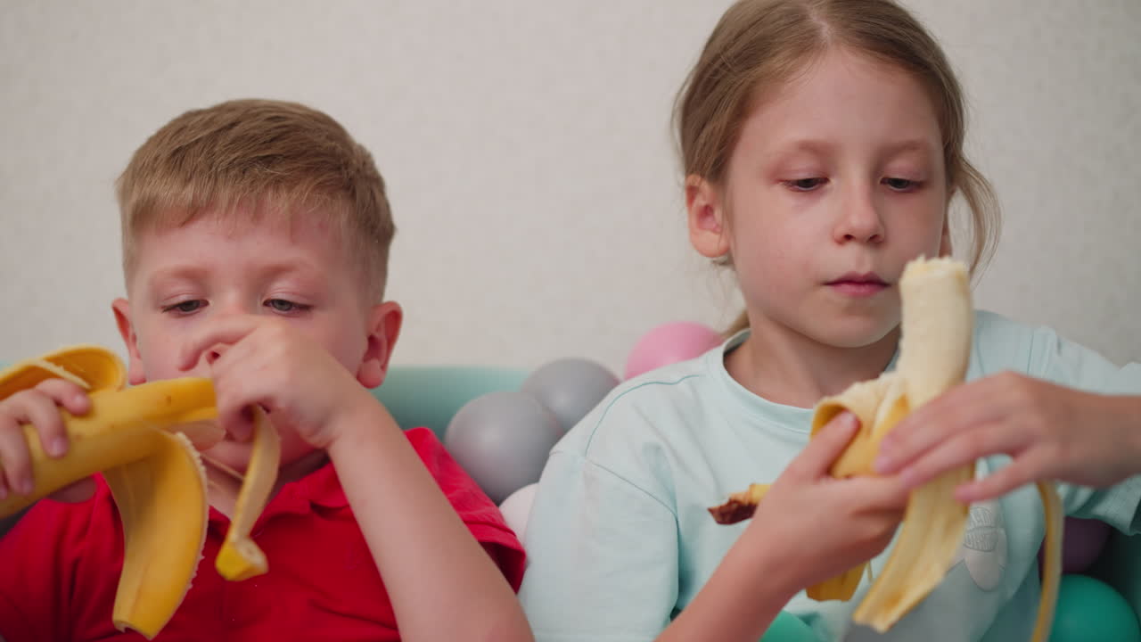 Brother and sister sitting in pastel ball pit peeling and eating bananas, boy wearing red shirt and girl in cyan top focused on fruit, natural candid moment with playful atmosphere
