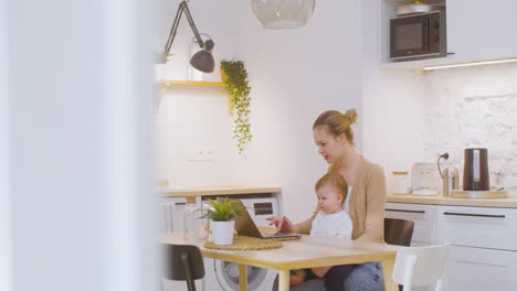 young woman working on laptop computer while sitting with baby boy at home