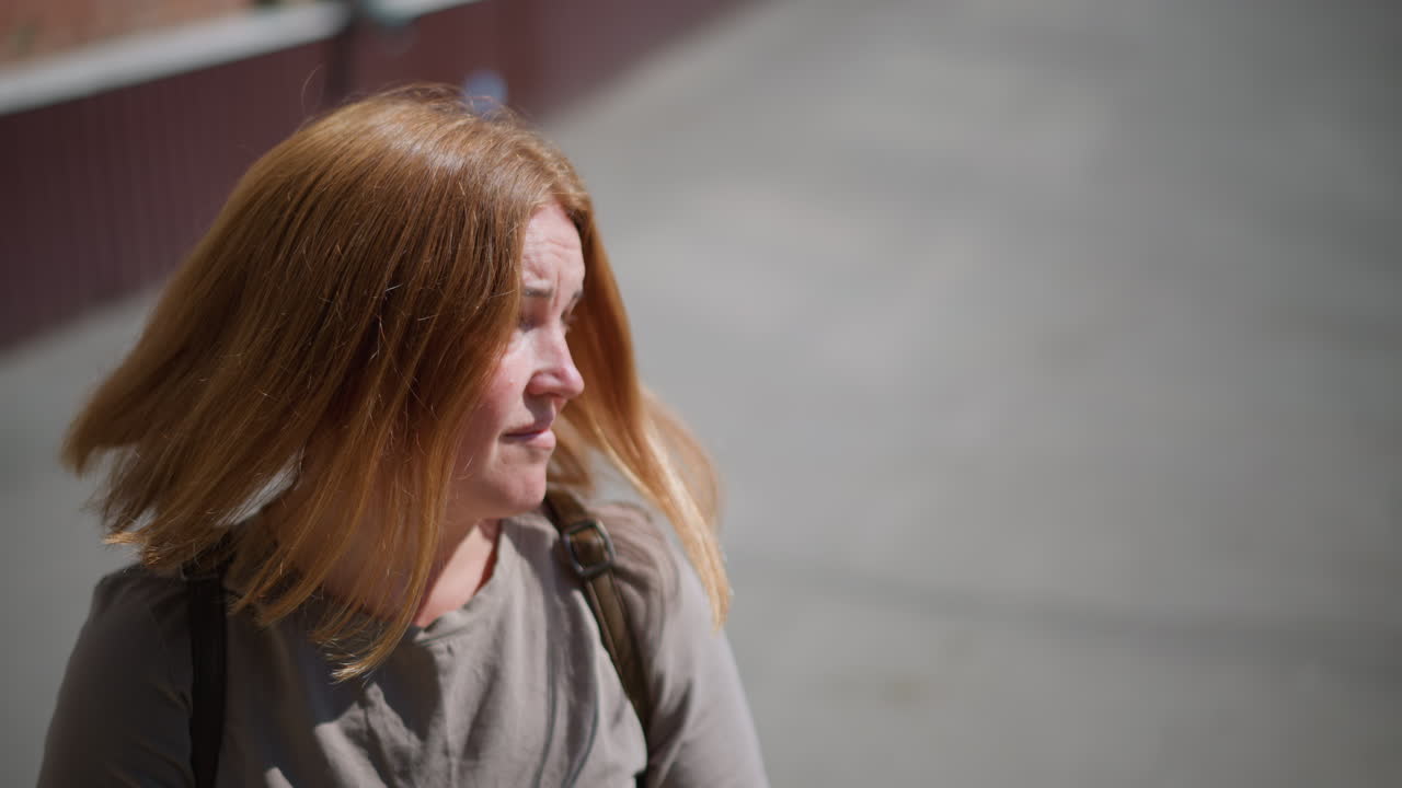 Aerial view of young woman standing under bright sunlight close to school building, looking around and checking time with fitness bracelet, showing sadness and quiet emotion on warm summer day