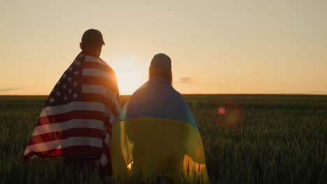 pareja con las banderas de ucrania y los ee.uu. de pie uno al lado del otro y mirar la puesta de sol sobre un campo de trigo