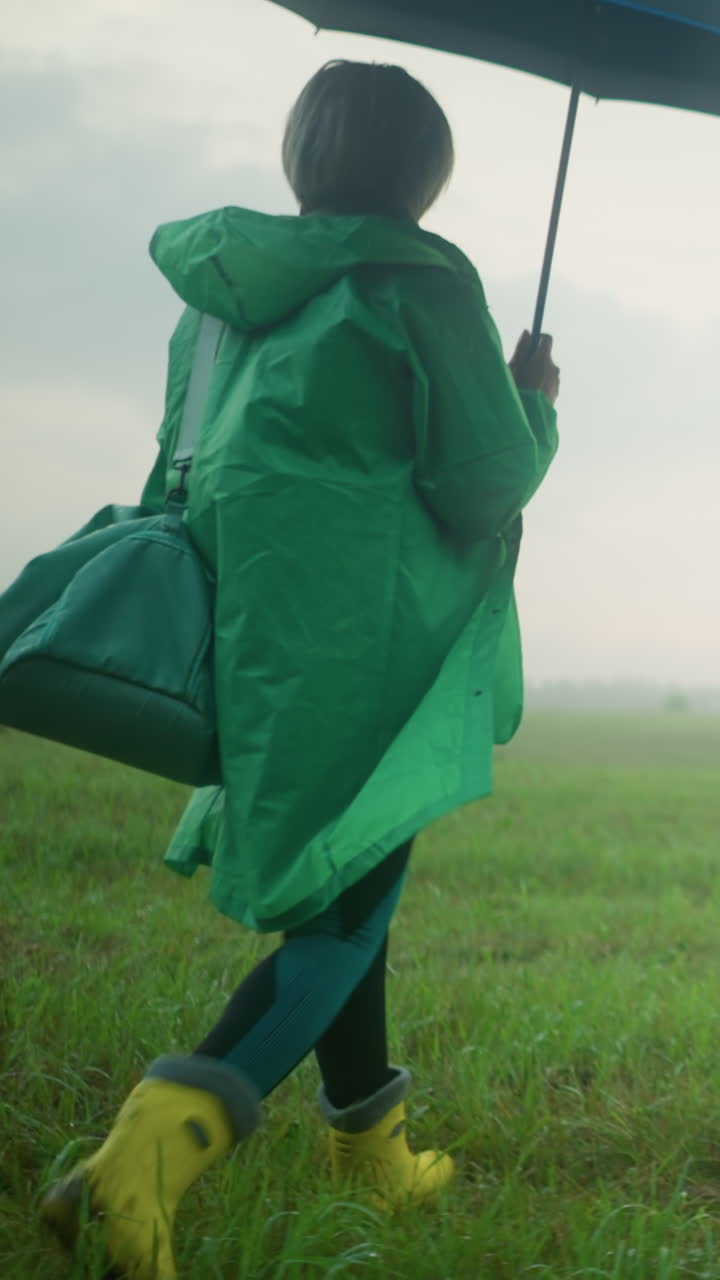 초록색 레인코트를 입은 여자가 안개가 인 초록색 을 고, 민트색 가방과 열린 우산을 들고, 넓은 풍경은 어져있는 관목, 나무, 그리고 배경에 구름이 인 하늘에 둘러싸여 있습니다.