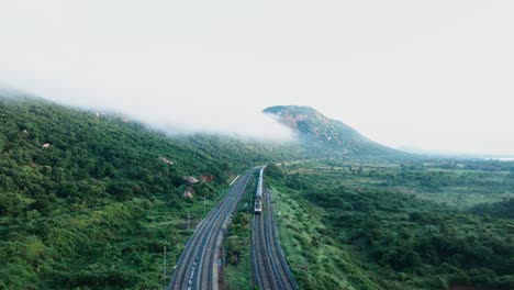 Aerial-backwards-shot-of-train-driving-in-green-Western-Ghats-of-India-during-cloudy-day