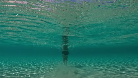 underwater back view in shallow water of man walking on seabed raising clouds of sand floating in turquoise tropical sea