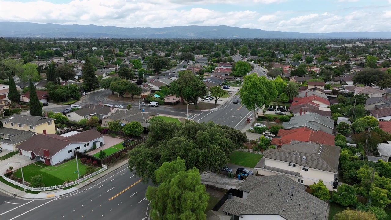 Bairro suburbano tranquilo com casas unifamiliares e telhados de telha vermelha ao longo de estradas largas que se cruzam, copa densa de árvores e cordilheira sob céu nublado. Califórnia, EUA. Imagem aérea descendente