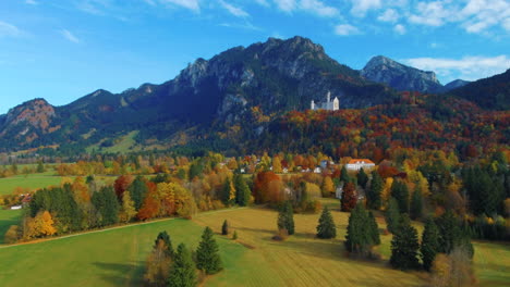 Angled-shot-of-drone-gliding-slowly-while-flying-over-village-houses-towards-castle-on-hill-over-scenic-autumn-field-in-the-afternoon-near-the-Neuschwanstein-Castle-in-Germany,-Europe,-wide-view