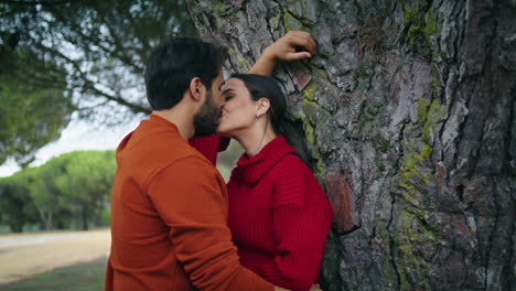 una pareja encantadora besándose en el parque apoyándose en un árbol de cerca. una pareja familiar de pie abrazándose