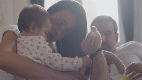 Feliz-Madre-En-La-Cama-Sosteniendo-Un-Oso-De-Peluche-Y-Jugando-Con-Su-Pequeña-Hija-Mientras-Papá-Los-Observa