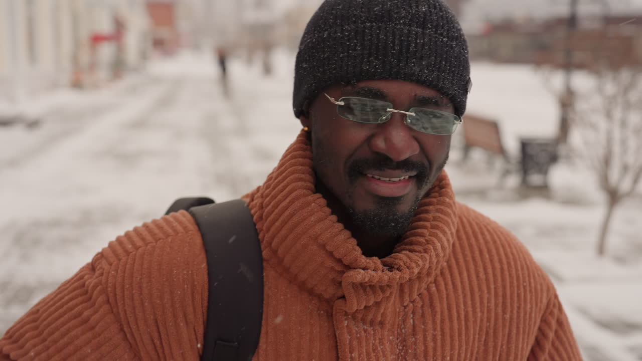Uomo di colore che cammina per una strada innevata sorridendo, maglione arancione accogliente, berretto lavorato a maglia, occhiali, zaino, neve che cade, passeggiata fiancheggiata da panchine, ritratti ravvicinati, espressione calda, pendolarismo urbano rilassato, passo tranquillo