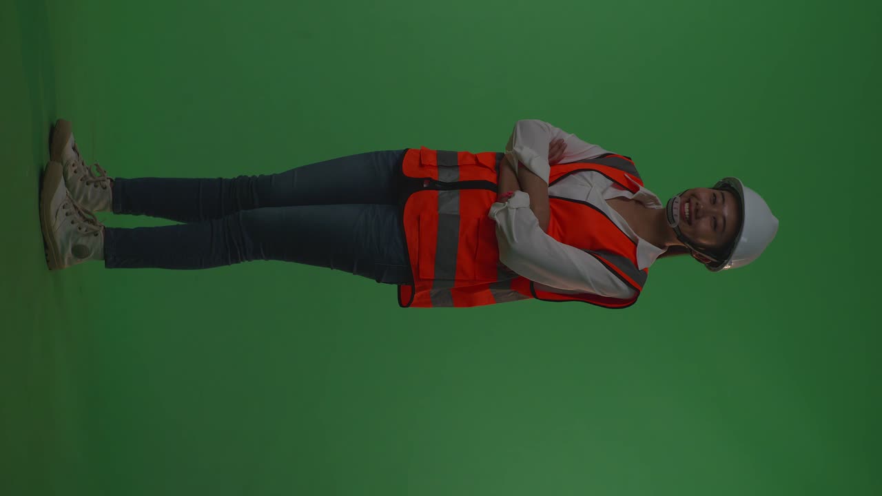 Full Body Of Asian Female Engineer With Safety Helmet Crossing Her Arms And Smiling To Camera While Standing In The Green Screen Background Studio, During Sunset Or Sunrise Time