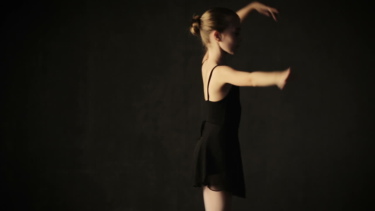 Little adorable young ballerina in tutu and pointe in the black studio