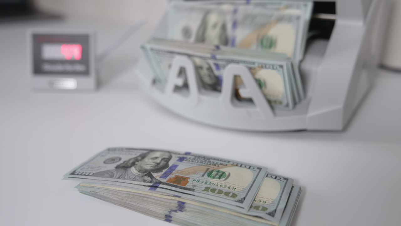 A wad of one hundred dollar bills lies on the table. Cash counting machine counts one hundred dollar bills at backdrop. Close up.