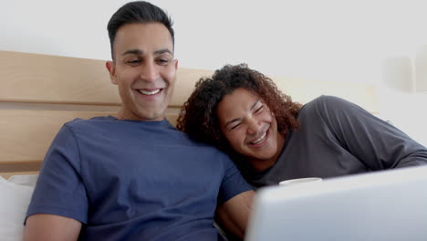 Happy-diverse-gay-male-couple-on-bed,-having-coffee-and-using-laptop-in-the-morning,-slow-motion