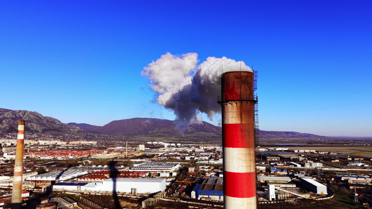 Ciminiera a strisce rosse e bianche che rilascia emissioni, con una città e una catena montuosa sullo sfondo. Primo piano aereo di una ciminiera industriale che emette fumo bianco denso.