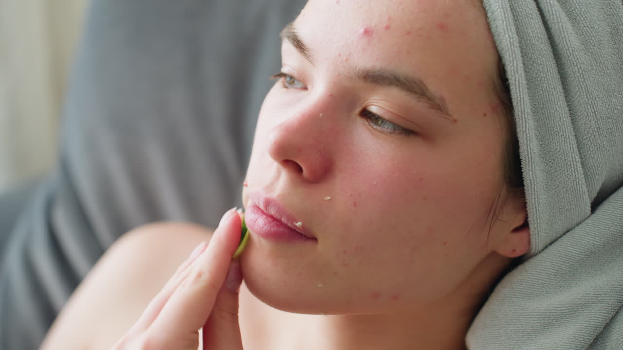 Woman with towel-wrapped hair relaxing on couch, gently massaging cheek with cucumber slice during soothing skincare routine, eyes closed in peaceful ambiance filled with soft natural lighting