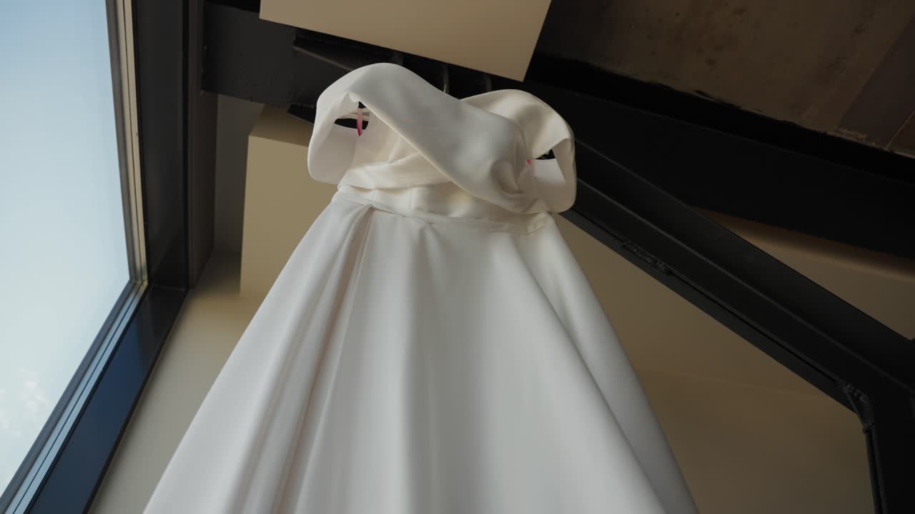 Close-up of a white satin wedding dress hanging indoors, showcasing its elegant neckline and soft flowing fabric, perfect for a romantic bridal moment, zooming in