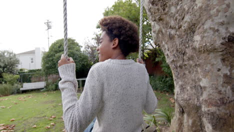 happy african american boy swinging on tree swing in garden, in slow motion