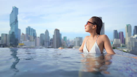 Junge-Asiatische-Frau,-Die-Sich-Im-Pool-Mit-Wolkenkratzern-Im-Hintergrund-Entspannt