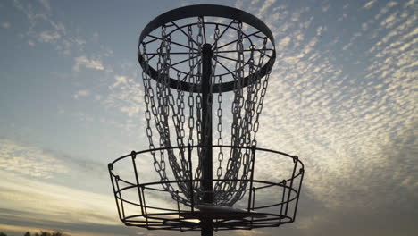 2 discs simultaneously go into disc golf basket that is silhouetted by the late evening sky with beautiful clouds