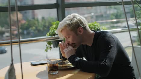 young man with blond hair enjoying a sandwich and iced coffee at a cafe table by a window with city views, natural light, and potted plants. relaxed and casual dining scene