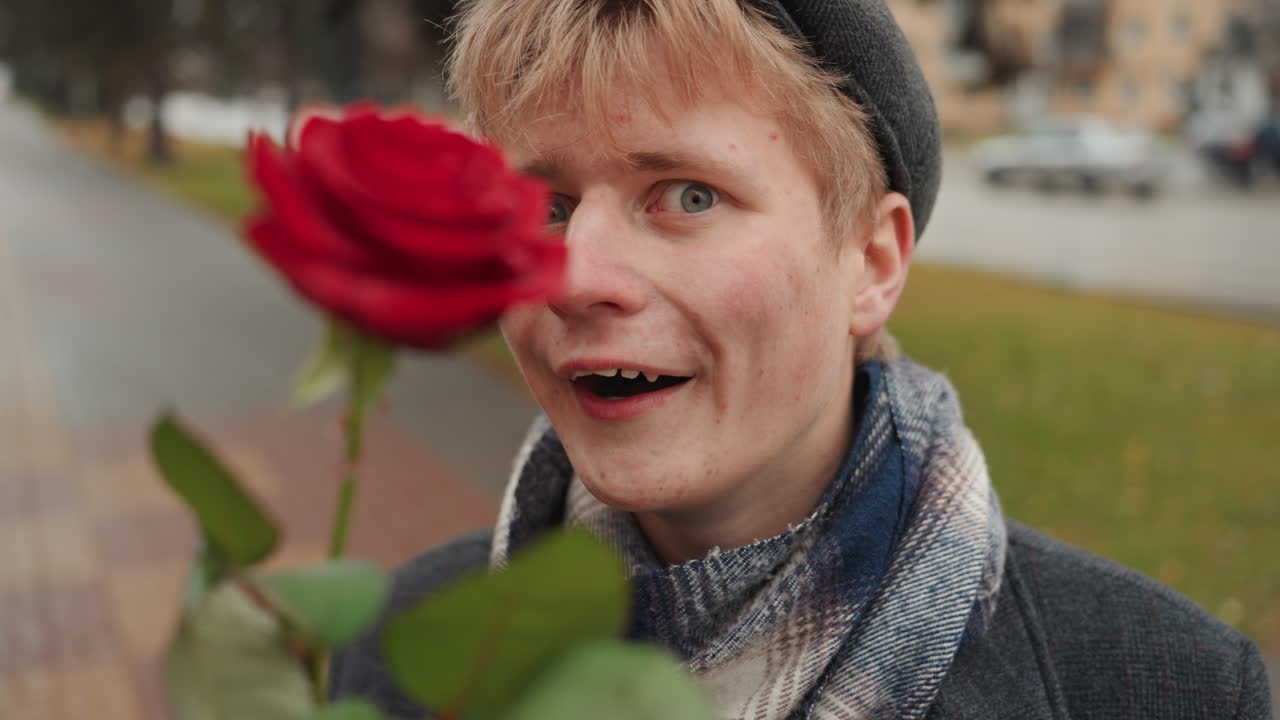 Creatore bianco sul marciapiede della città che presenta una rosa, espressione giocosa e sorriso civettuolo, berretto e sciarpa accogliente, ritratto ravvicinato che mostra il fiore rosso e il fascino genuino, atmosfera da poeta di strada e spontanea