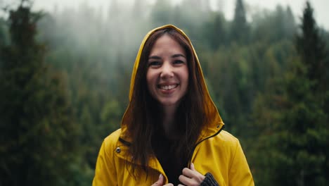 a happy girl in a yellow jacket with a hood stands against the backdrop of a coniferous mountain forest, she smiles and looks at the camera