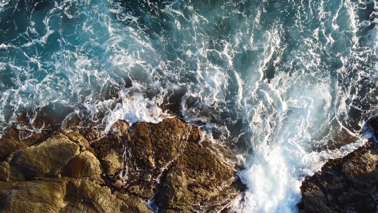 Vue aérienne plongeante de vagues s'écrasant contre des rochers, en arrière-plan.