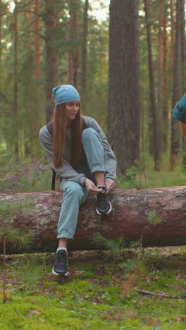 siblings take a break in serene forest surroundings, one sitting on a fallen tree, tying her shoe with a blue bandana on her head, while her companion stands close, engaging in conversation