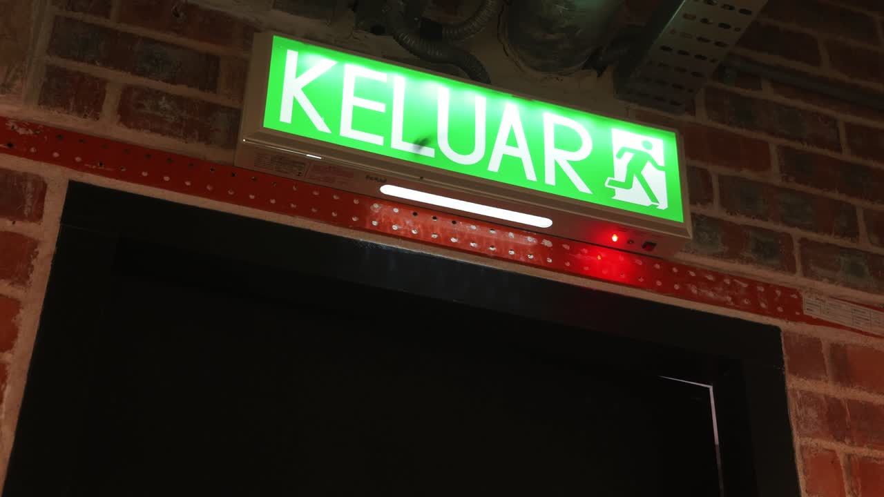A bright green KELUAR Exit sign illuminated above a dark fire door opening, mounted below industrial cable trays and against a red brick wall