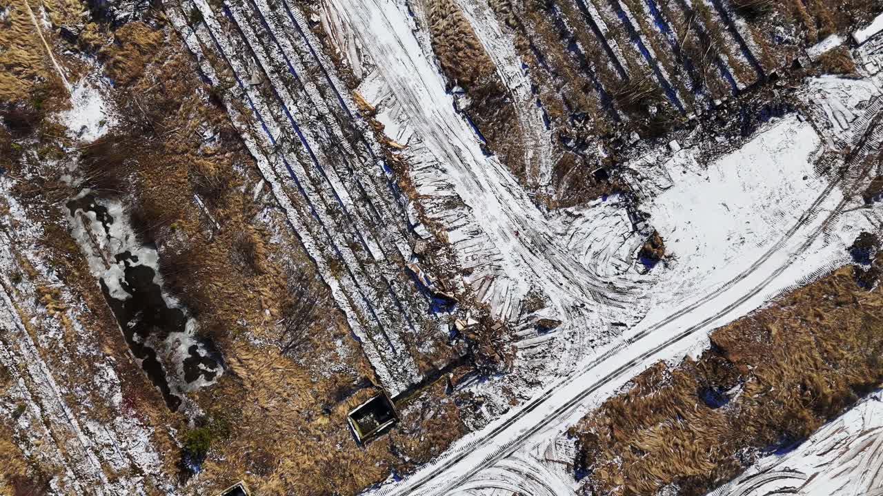 An expansive construction area on the outskirts of a town, with muddy and snowy terrain, visible machinery, and nearby warehouses and residential buildings.