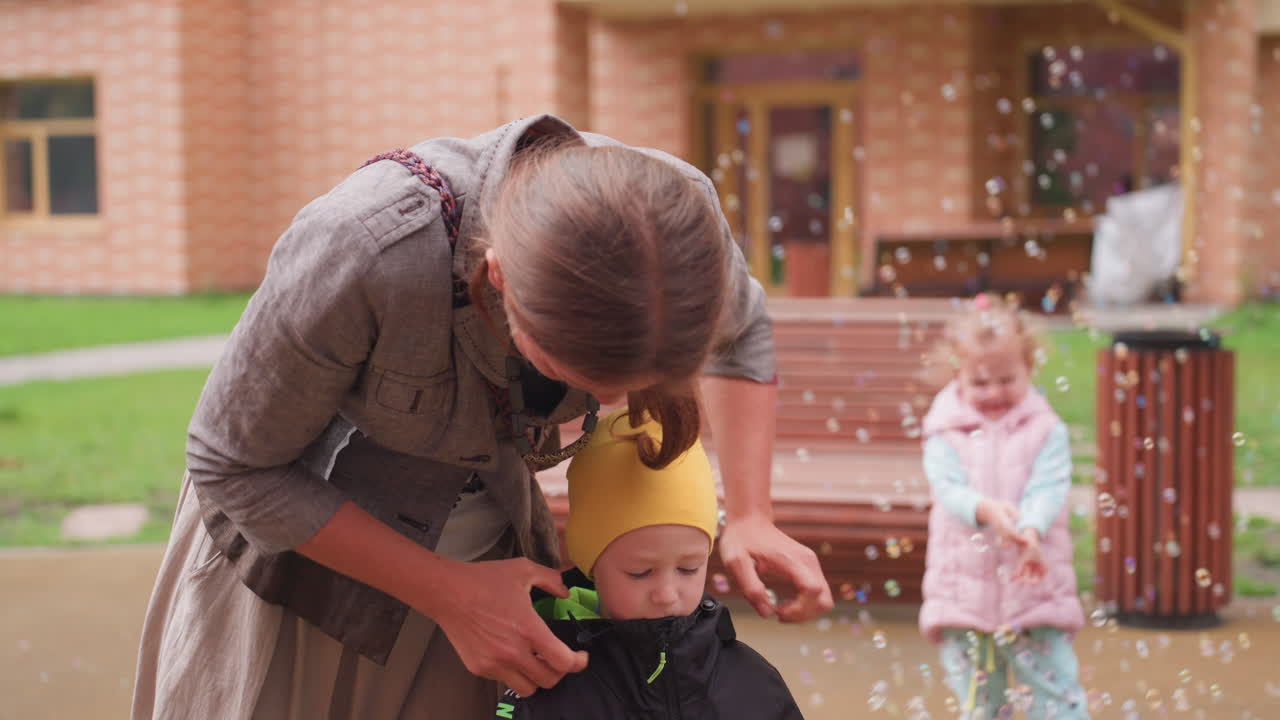 Caring mother bends down to zip son jacket as little girl joyfully plays among floating bubbles in bright outdoor courtyard, expressing warmth, care, and playful family moment