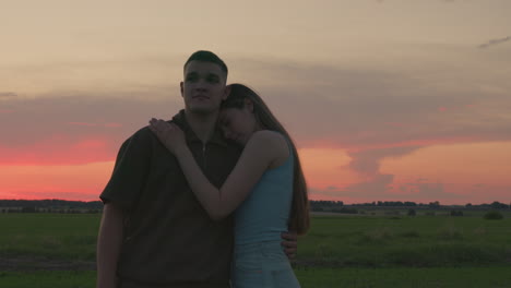 young couple embraces in serene field during glowing sunset as woman holds man closely with calm expressions, surrounded by warm light and tranquil atmosphere