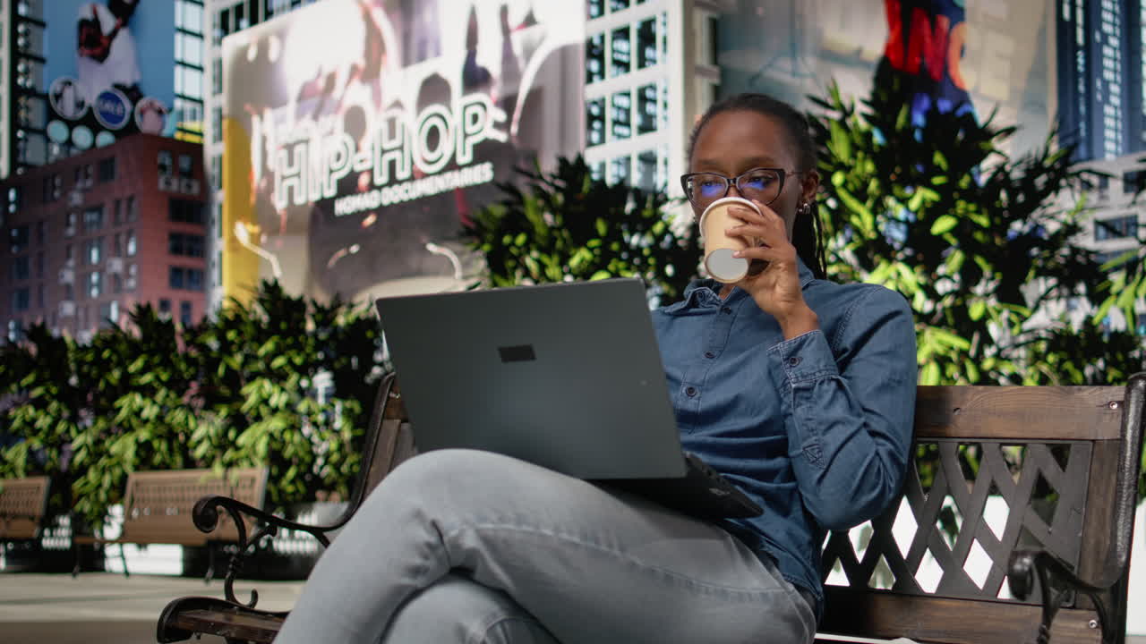 Digital nomad drinking coffee working with laptop on city park bench