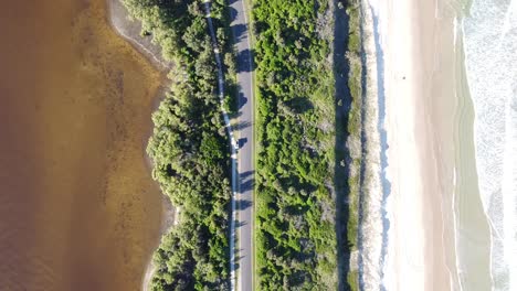 Vista-Aérea-De-Un-Dron-De-Un-Coche-Que-Va-Por-Una-Carretera-Vacía-Entre-La-Playa-Y-Un-Lago-En-Australia