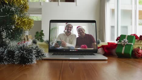 Smiling-biracial-father-and-son-wearing-santa-hats-on-christmas-video-call-on-laptop