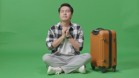 full body of asian male traveler with luggage praying for something while sitting in the green screen background studio