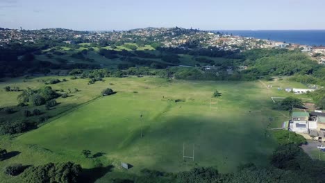 Drone-flying-over-a-rugby-soccer-field-overlooking-Bluff-golf-course-with-green-scenery-with-a-road-dividing-and-the-ocean-in-the-background