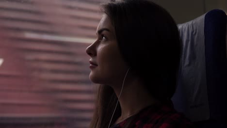 Pensive-girl-travels-by-train.-Sitting-next-to-the-window.-Looking-up.-Listen-music-in-earphones.-Thoughtful-look.-Side-view