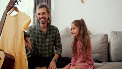 un padre solitario, un hombre moreno en una camisa a cuadros verde, se comunica con su hija y vaporiza un vestido amarillo usando un vaporizador mientras está sentado en un sofá gris en un apartamento moderno