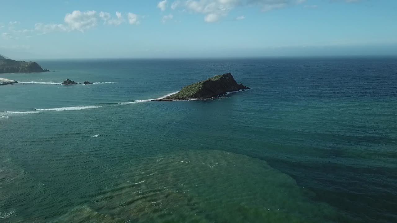 A scenic shot taken from the open Mediterranean Sea, looking towards the coast. A small rocky islet sits very close to the sandy shoreline