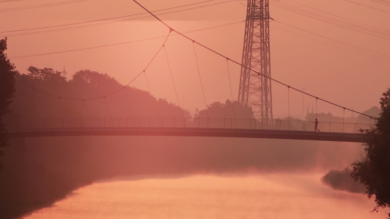 Silhoutte of jogger crosses urban bridge over misty river at fiery orange dawn