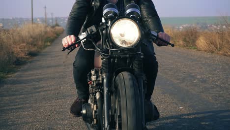 front view of a unrecognizable man in black leather jacket riding motorcycle on a country road holding a handlebar. big headlight turned on