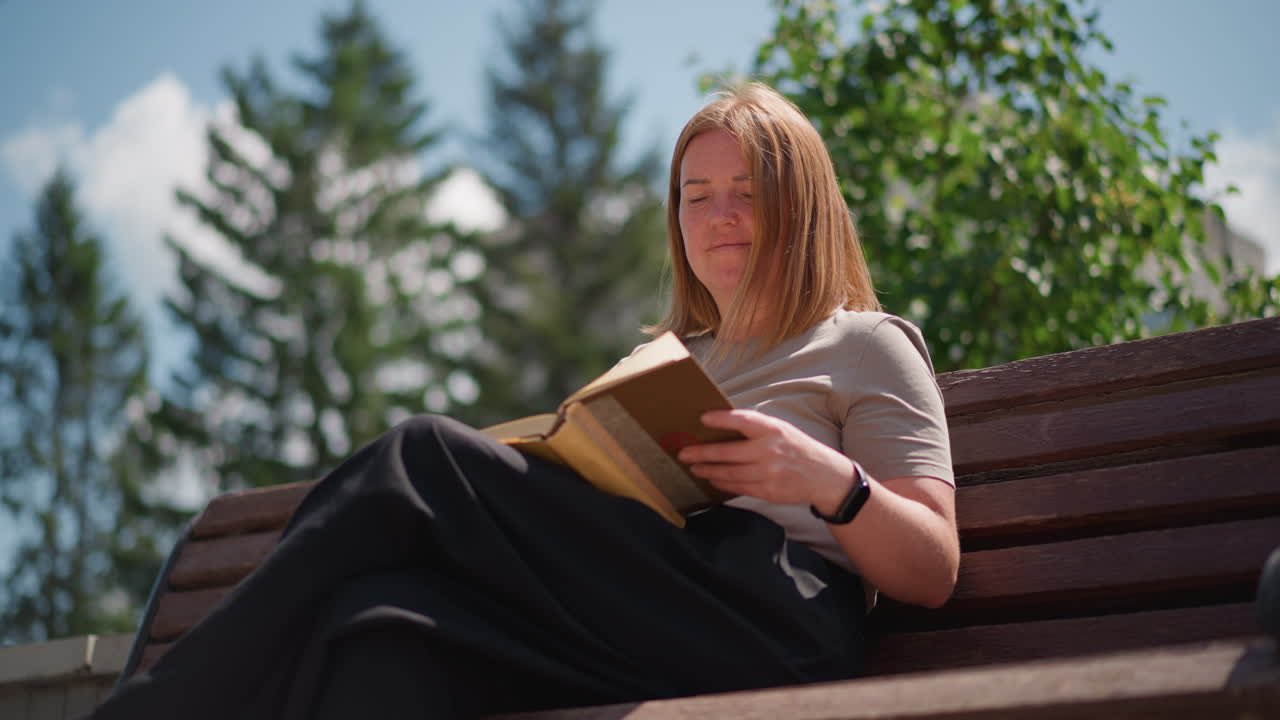 Pupil sitting on wooden bench under sunlight closes book and presses it to chest while turning to watch passing cars and pigeon nearby in urban park peaceful outdoor scene
