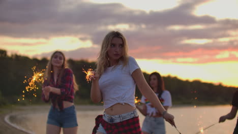 the blonde female with nude waist and slim figure is dancing with big bengal lights in her hands on the sand beach with her friends. this is a nice summer evening on the open air party.