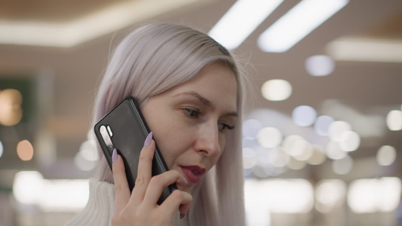 close up of stylish woman with lilac hair in white sweater holding smartphone to ear listens intently in busy mall environment overhead lights and blurred shoppers behind create dynamic retail scene
