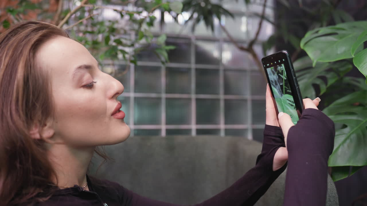 Close-up of woman with blond hair and soft smile holding smartphone to take picture of green leaf, focused and immersed in snapping shot, seated near indoor plants with natural lighting